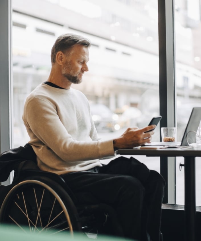 A man in a wheelchair sits in a café window while looking at his cellphone and worrying about romance scams.